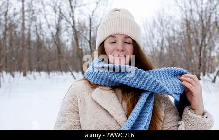 Jeune femme souriante avec les yeux fermés portant une écharpe bleue dans la forêt d'hiver Banque D'Images