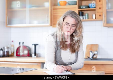 Femme mature sérieuse écrivant sur papier dans la cuisine à la maison Banque D'Images