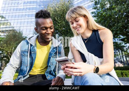 Amis multiraciaux souriants partageant un téléphone intelligent assis près du bâtiment Banque D'Images