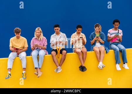 Amis heureux avec des vêtements colorés assis sur jaune à l'aide de leurs smartphones Banque D'Images