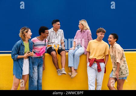 Groupe d'amis amis avec des vêtements colorés assis sur le mur jaune discutant ensemble Banque D'Images