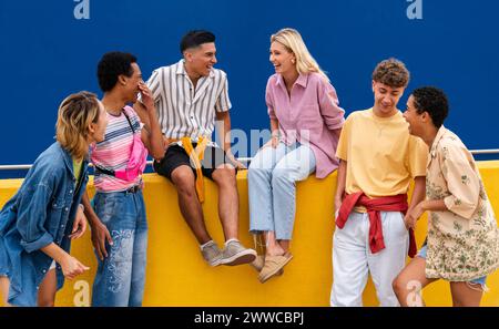 Groupe d'amis amis avec des vêtements colorés assis sur le mur jaune discutant ensemble Banque D'Images