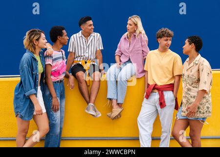 Groupe d'amis amis avec des vêtements colorés assis sur le mur jaune discutant ensemble Banque D'Images