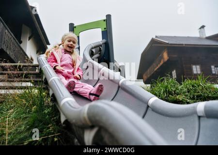 Fille en imperméable rose glissant vers le bas toboggan à l'aire de jeux Banque D'Images