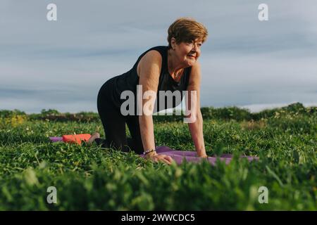 Femme senior pratiquant le yoga sur tapis d'exercice au milieu des plantes à la prairie Banque D'Images
