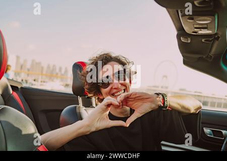 Femme heureuse portant des lunettes de soleil montrant la forme de coeur assis dans la voiture convertible le jour ensoleillé Banque D'Images