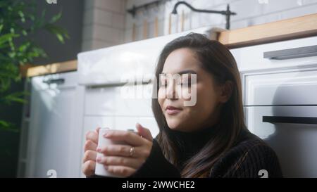 Détendue jeune femme asiatique assise sur le sol dans la cuisine, buvant du café du matin ou du thé chaud, profitant de passer du temps dans sa propre maison. Banque D'Images