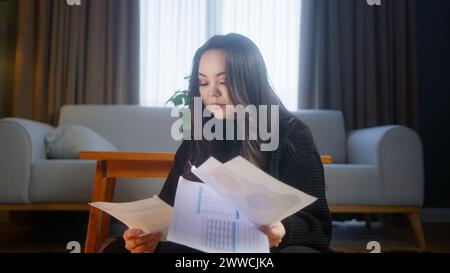 Fille asiatique ennuyée assise sur le sol à la maison tenant et regardant des papiers, étudiant. Banque D'Images