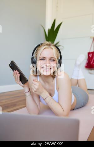 Plan vertical de jeune femme blonde dans des écouteurs, couchée sur un tapis en caoutchouc, à l'aide d'un ordinateur portable, tenant un téléphone portable, écoute de la musique ou un podcast d'entraînement, faisant Banque D'Images