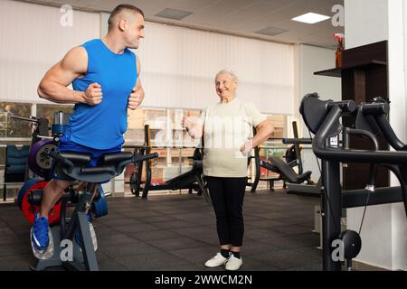 Un beau jeune entraîneur montre à la vieille femme comment faire l'exercice sur le simulateur. Les gens sourient, sont heureux, montrent la classe, le pouce. Super. Sport actif Banque D'Images