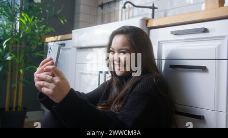 Fille asiatique ravie de sourire et de prendre selfie avec son téléphone portable tout en étant assis sur le sol dans la cuisine à la maison Banque D'Images