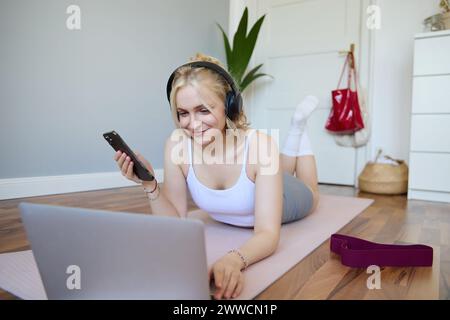 Portrait de l'entraînement de jeune femme, regarder des vidéos d'exercice sur ordinateur portable dans des écouteurs, couché sur tapis en caoutchouc avec téléphone portable et souriant Banque D'Images