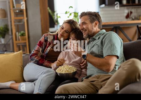 Une famille de trois est confortablement nichée sur un canapé, leurs visages reflétant l'excitation et l'attention comme ils partagent un bol de pop-corn pendant un susp Banque D'Images