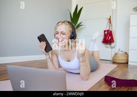 Portrait de l'entraînement de jeune femme, regarder des vidéos d'exercice sur ordinateur portable dans des écouteurs, couché sur tapis en caoutchouc avec téléphone portable et souriant Banque D'Images