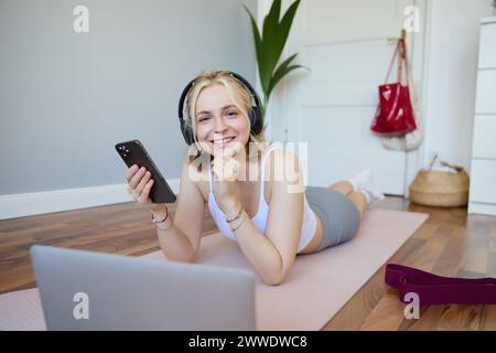 Portrait de l'entraînement de jeune femme, regarder des vidéos d'exercice sur ordinateur portable dans des écouteurs, couché sur tapis en caoutchouc avec téléphone portable et souriant Banque D'Images