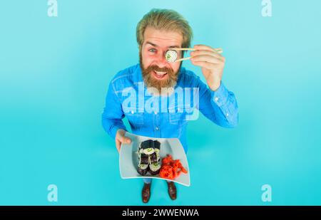 Homme barbu souriant dans une chemise décontractée en denim couvrant l'oeil avec rouleau de sushi. Cuisine japonaise traditionnelle. Beau mâle avec assiette de délicieux sushis maki Banque D'Images