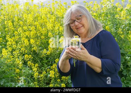 Femme mature souriante dans la soixantaine avec des cheveux blancs et des lunettes tenant un bouquet de marguerites dans ses mains avec un champ avec des fleurs en arrière-plan Banque D'Images