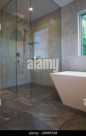 Cabine de douche en verre transparent et baignoire blanche autoportante en forme de récipient dans la salle de bains avec carrelage en céramique grise et murs au rez-de-chaussée à l'intérieur Banque D'Images