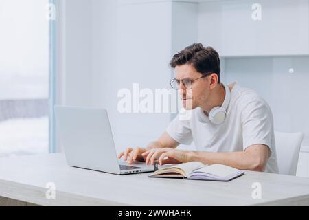Un homme avec des écouteurs travaille intensément sur un ordinateur portable dans un bureau à domicile lumineux et minimaliste, incarnant l'expérience de télétravail moderne. Télétravail concentré Banque D'Images