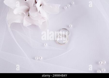 Fond de mariage chic dans les tons blancs. Deux bagues de fiançailles en platine, une bague en diamant sur un voile de mariée blanc classique avec des fleurs fraîches. Vue de dessus Banque D'Images