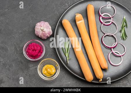 Trois longues saucisses, brins de romarin et rondelles d'oignon sur plaque noire. Moutarde et ail, piment sur table. Fond noir. Pose à plat Banque D'Images