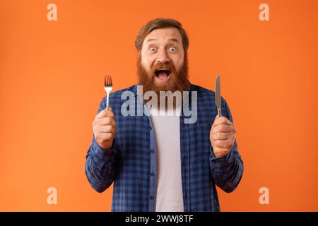 Homme barbu affamé tient couteau et fourchette exprimant l'excitation, studio Banque D'Images