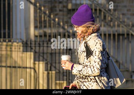 Une jeune femme marche avec une tasse jetable de boisson chaude dans la rue et éclairée par le soleil levant. Dublin, Irlande. Banque D'Images