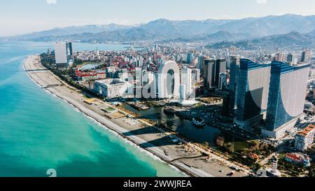 La photo aérienne capture un paysage urbain moderne le long de la côte, avec des gratte-ciel saisissants juxtaposés à la mer bleue sereine et encadrés par des montagnes majestueuses. Géorgie, Batoumi, février 2023. Banque D'Images