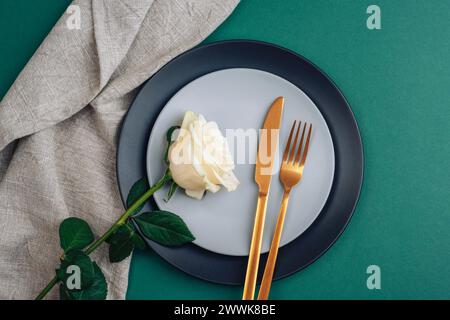 Table place avec fleur de rose blanche, assiettes et couverts dorés sur fond vert foncé. Vue de dessus, pose à plat. Banque D'Images