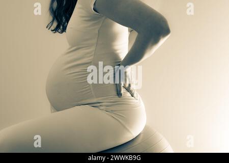 Description : femme enceinte avec bosse de bébé traite les maux de dos et à l'aide d'un ballon de gymnastique. Dernier mois de grossesse - semaine 36. Vue latérale. Dos blanc Banque D'Images