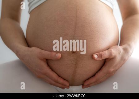 Description : gros plan de la femme assise et tenant doucement son ventre de bébé enceinte très rond. Vue de face. Fond blanc. Prise de vue brillante. Banque D'Images