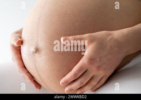 Description : gros plan d'une mère assise et tenant doucement son ventre de bébé enceinte très rond. Vue latérale. Fond blanc. Prise de vue brillante. Banque D'Images