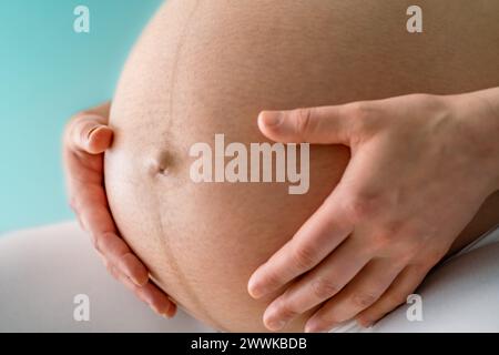 Description : gros plan d'une femme assise tenant doucement sa bosse de bébé enceinte très ronde. Vue latérale. Fond turquoise. Prise de vue brillante. Noir an Banque D'Images