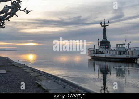 Beschreibung : Die Imperia-Statue und die traditionelle Autofähre an der Hafeneinfahrt mit ruhigem See und den Alpen am Horizont in der Morgendämmerung Banque D'Images