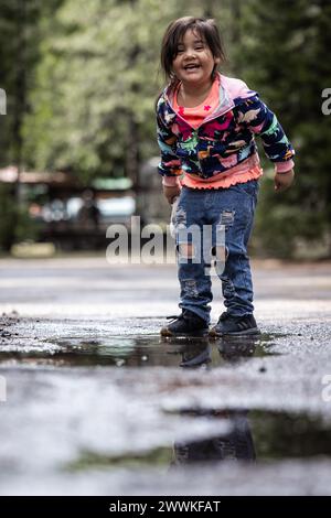 Une jeune fille se tient debout dans une flaque d'eau, portant une veste bleue et rose. Elle sourit et elle s'amuse Banque D'Images