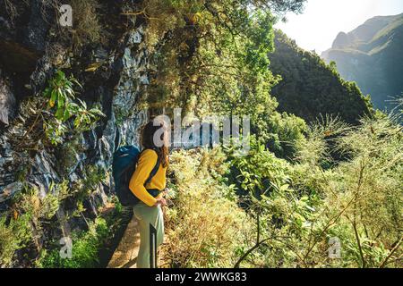 Description: Femme touristique bénéficiant d'une vue depuis le dessous d'un grand mur de roche le long du canal d'eau à la falaise abrupte à travers la forêt tropicale de Madère. Levada de Caldeir Banque D'Images