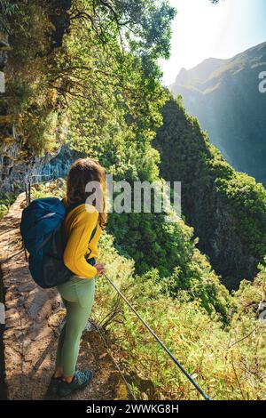 Description: Femme touristique bénéficiant d'une vue depuis le dessous d'un grand mur de roche le long du canal d'eau à la falaise abrupte à travers la forêt tropicale de Madère. Levada de Caldeir Banque D'Images