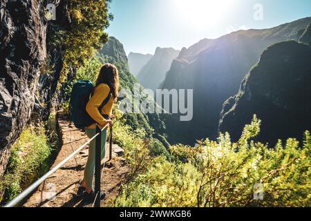 Description: Backpacker femme jouissant d'une vue panoramique depuis le dessous grand mur de roche le long du canal d'eau à la falaise raide à travers la forêt tropicale de Madère. Levada Banque D'Images