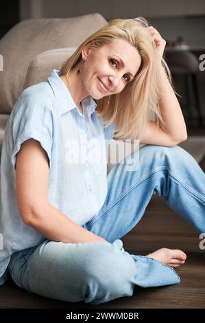 Portrait de jolie femme avec les cheveux blonds relaxant à côté du canapé. Belle femme assise sur plancher en bois. Banque D'Images