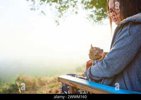 Vue latérale de la femme asiatique assise sur le banc avec son chaton pendant la matinée brumeuse dans la forêt Banque D'Images