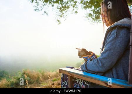 Vue latérale de la femme asiatique assise sur le banc avec son chaton pendant la matinée brumeuse dans la forêt Banque D'Images