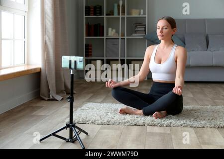 femme faisant du yoga à la maison sur le tapis, assise en position lotus avec son téléphone et trépied pour enregistrer des vidéos ou en streaming en direct pour un fitne en ligne Banque D'Images