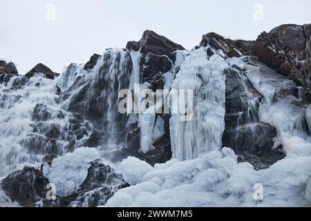 Une cascade partiellement gelée coule au milieu des formations de glace sur une falaise accidentée pendant une froide journée d'hiver Banque D'Images