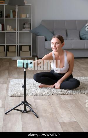 femme assise sur le sol dans un pantalon de yoga enregistrant une vidéo de fitness en ligne avec son téléphone et son trépied, souriant à la caméra dans un cadre de salon Banque D'Images