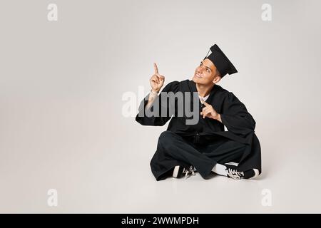 jeune homme joyeux dans la robe de troisième cycle et casquette assis et montrant avec les doigts vers le haut Banque D'Images
