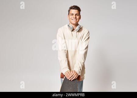 étudiant joyeux en sweat à capuche blanc et écouteurs appuyés sur le skateboard sur fond clair Banque D'Images