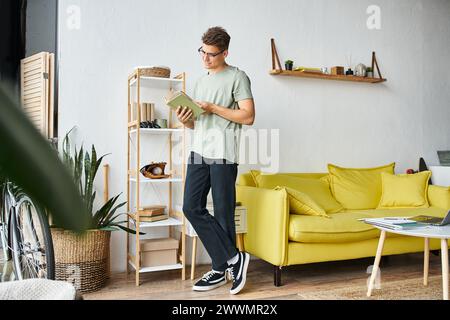 bel étudiant dans les années 20 avec des cheveux bruns et des lunettes de vision dans le salon debout et livre de lecture Banque D'Images