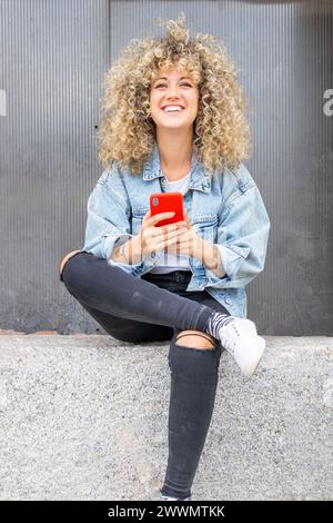jeune femme caucasienne aux cheveux blonds bouclés, souriante , discutant avec son téléphone intelligent rouge Banque D'Images
