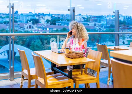 Portrait d'une jeune femme caucasienne souriante aux cheveux blonds bouclés, travaillant dans un café avec son ordinateur portable et son téléphone intelligent Banque D'Images