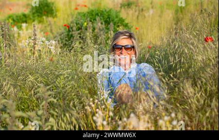 fröhliche Junge Frau mit Sonnenbrille sitzt in einer Blumenwiese *** jeune femme joyeuse avec des lunettes de soleil est assise dans un pré de fleurs Banque D'Images